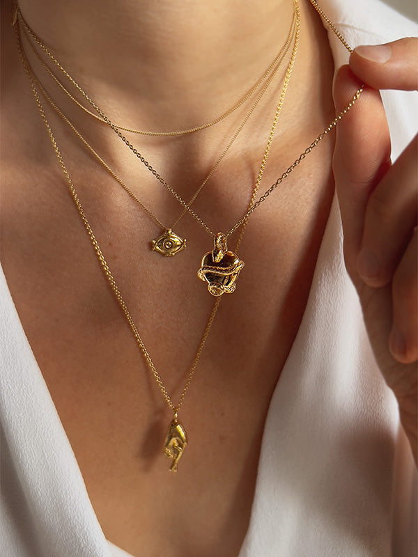 gold charm necklaces on a model's neck. She is holding the chain of one necklace. Charms are a snake around a heart for wisdom, and eye for protection and a hand with fingers crossed for good luck